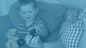 A young boy with a hearing aid sits on a couch and hands a toy to an adult.